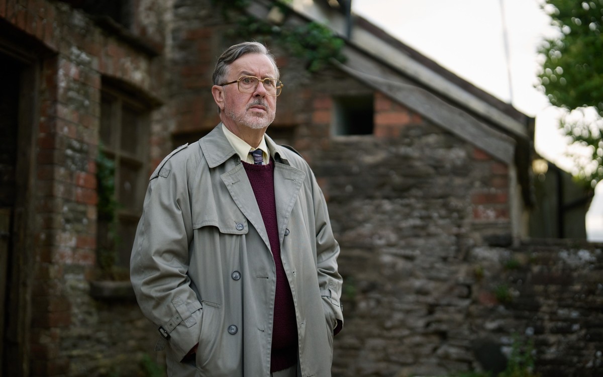 BBC confirms The Witch Farm release date 5 Jeremy Swift as Eddie Burks stands outside a brick and timber building, wearing glasses and a raincoat.
